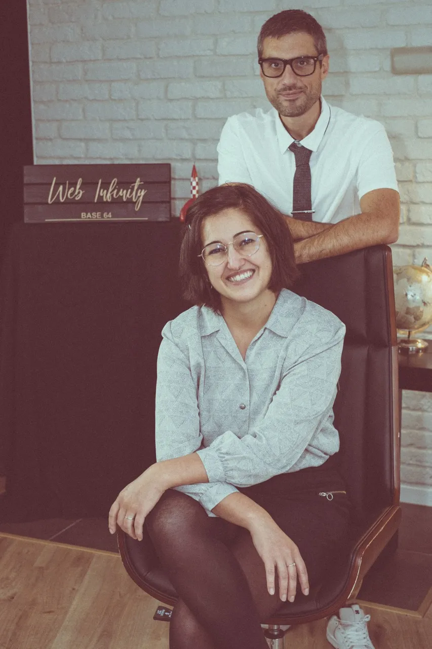 Noélène assise et souriante, Alexandre derrière elle, sous l'enseigne Web-Infinity de l'agence